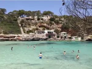 Personas bañándose en las aguas cristalinas de color turquesa de Cala s'Almunia, en Mallorca, rodeadas de acantilados rocosos, antiguas casetas de pescadores y pinos mediterráneos en un soleado día de verano.