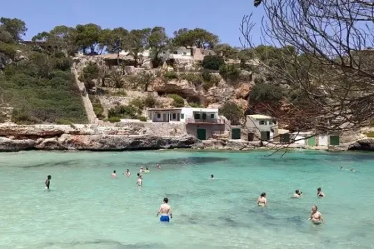 Personas bañándose en las aguas cristalinas de color turquesa de Cala s'Almunia, en Mallorca, rodeadas de acantilados rocosos, antiguas casetas de pescadores y pinos mediterráneos en un soleado día de verano.