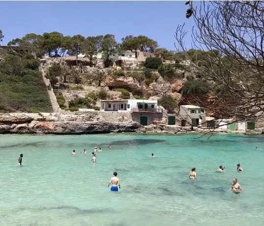 Personas bañándose en las aguas cristalinas de color turquesa de Cala s'Almunia, en Mallorca, rodeadas de acantilados rocosos, antiguas casetas de pescadores y pinos mediterráneos en un soleado día de verano.
