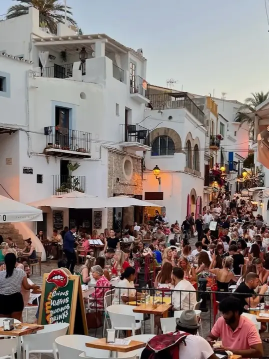 Animado atardecer en el casco antiguo de Ibiza (Dalt Vila), con terrazas de restaurantes, edificios encalados, calles empedradas y personas disfrutando de comida y bebida al aire libre.