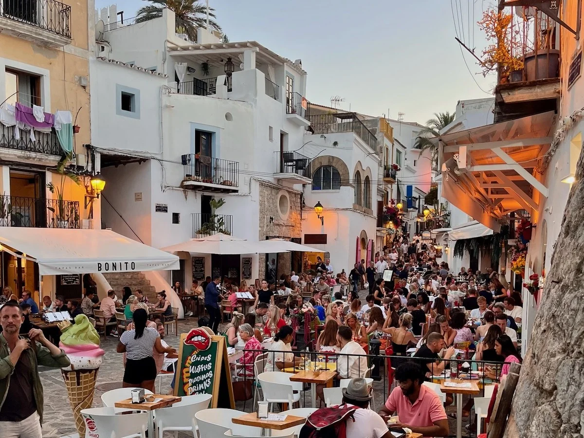 Animado atardecer en el casco antiguo de Ibiza (Dalt Vila), con terrazas de restaurantes, edificios encalados, calles empedradas y personas disfrutando de comida y bebida al aire libre.