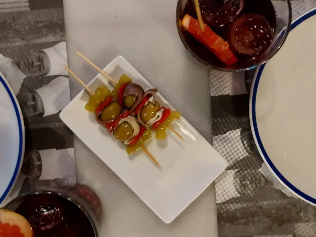 Pinchos de tapas estilo Gildas con anchoas, aceitunas y pimientos servidas junto a copas de vermut rojo en una mesa.