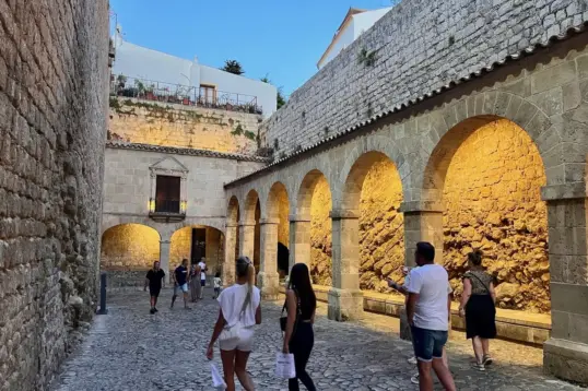 Visitors walking through the illuminated stone arches and cobbled streets of Dalt Vila, Ibiza's UNESCO-listed Old Town, during the early evening.