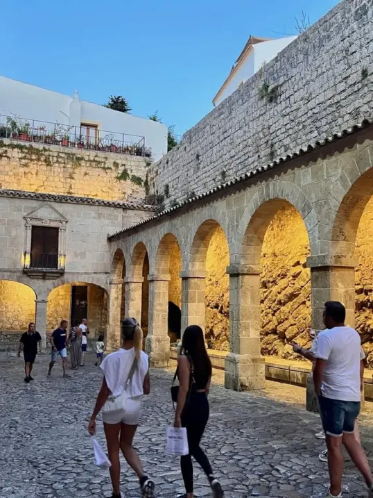 Visitors walking through the illuminated stone arches and cobbled streets of Dalt Vila, Ibiza's UNESCO-listed Old Town, during the early evening.