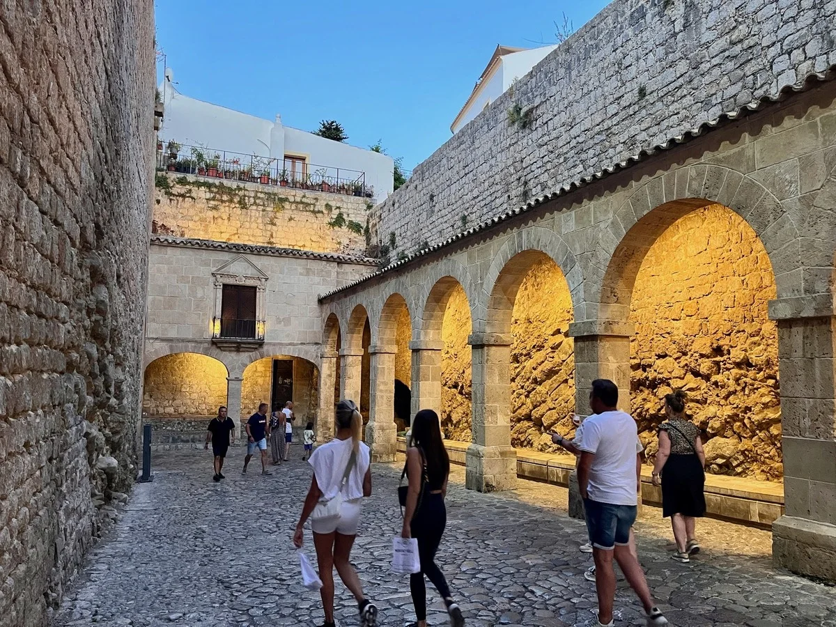 Visitors walking through the illuminated stone arches and cobbled streets of Dalt Vila, Ibiza's UNESCO-listed Old Town, during the early evening.