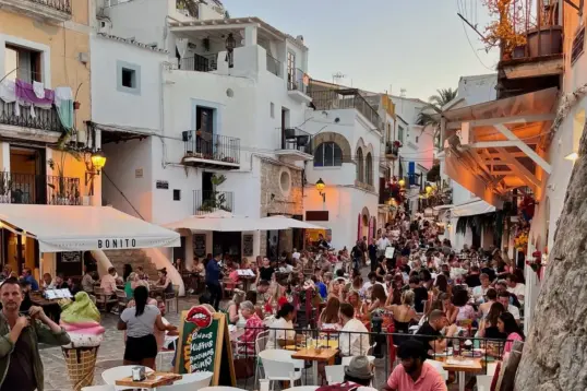Busy evening in Ibiza Old Town (Dalt Vila) with outdoor restaurant terraces, whitewashed buildings, cobbled streets, and diners enjoying food and drinks at sunset.